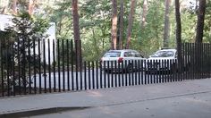 two cars parked in front of a black and white fence with trees behind it on the street