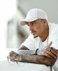 a man with tattoos on his arms and arm, leaning against a white wall looking at the camera