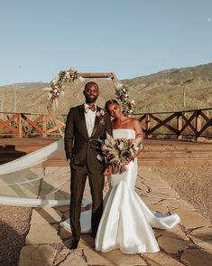 a man and woman standing next to each other in front of a wooden arch with flowers on it