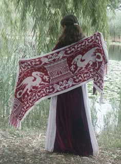 a woman holding a large red and white scarf in front of a lake with willow trees