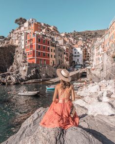 a woman sitting on top of a rock next to the ocean with buildings in the background