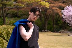 a young man wearing glasses and a blue cape standing in front of trees with pink flowers