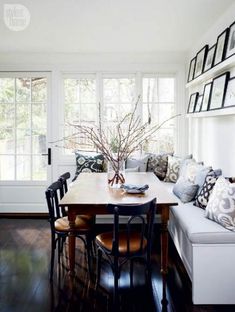 a dining room table surrounded by chairs in front of two windows with white walls and wooden flooring