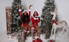 two women dressed as santa clause sitting in front of christmas trees