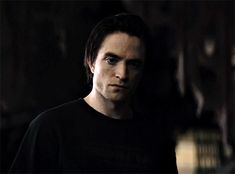 a man with dark hair and black shirt looking at the camera while standing in a dimly lit room