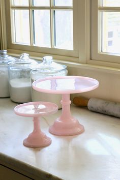 two pink cake plates sitting on top of a counter next to some jars and containers