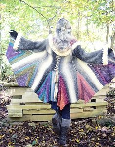 a woman is standing in the woods with her arms spread out, wearing a multicolored sweater and boots