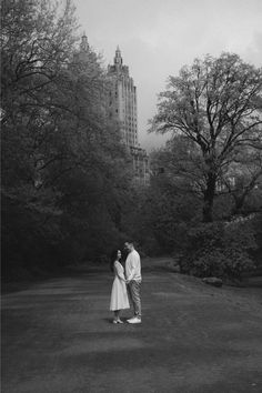 a man and woman standing next to each other in the middle of a park with trees