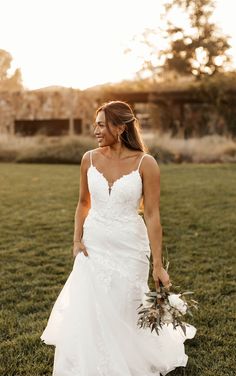 a woman in a white wedding dress holding a bouquet and walking through the grass at sunset