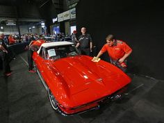 two men in red shirts and black pants standing next to a red car at an auto show