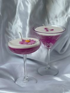 two martini glasses filled with purple liquid and topped with pink flowers on a white table cloth