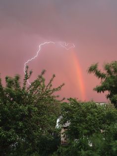 a rainbow appears in the sky over some trees and buildings as a lightning strikes behind it