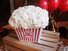 a popcorn box filled with white flowers on top of a wooden table next to balloons