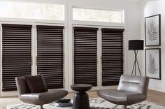 a living room with two chairs, a table and three windows that have blinds on them