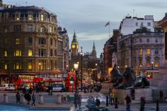 people are walking around in the city at dusk, with red double decker buses on the street