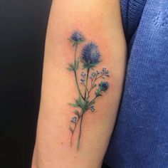 a small blue flower tattoo on the left upper half of the arm, with green leaves and purple flowers