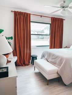 a white bed sitting next to a window in a room with orange drapes on the windowsill