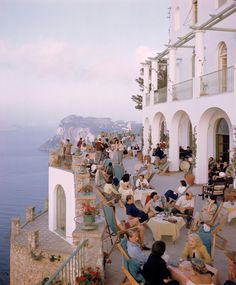 many people are sitting at tables outside by the water and some have drinks in their hands