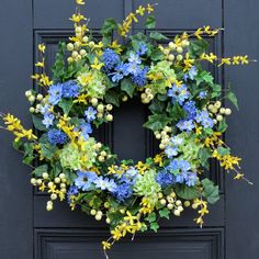 a blue wreath with yellow and green flowers hangs on a black front door, surrounded by greenery