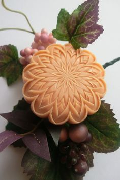 an orange soap with grapes and leaves around it on a white table top next to purple flowers