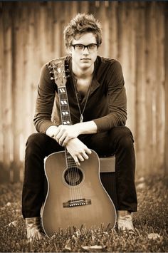 a man sitting in the grass with a guitar and bible verse on his back ground