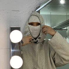 a woman wearing a hoodie and covering her face with a mask in front of a mirror