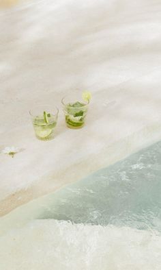 two glasses filled with cucumber slices sitting on top of a sandy beach next to the ocean