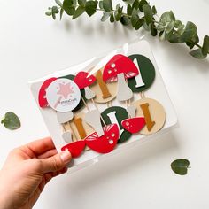 someone holding up some cupcake toppers in a box on a table with greenery