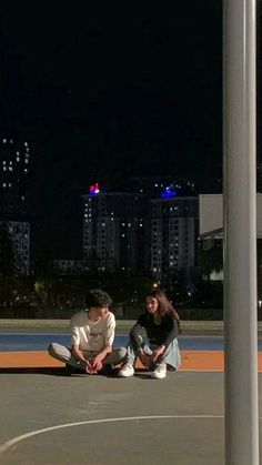 two people sitting on the ground in front of a basketball court with buildings in the background