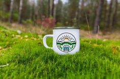 a white coffee cup sitting on top of a green grass covered field with trees in the background