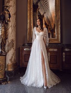 a woman standing in front of a painting wearing a white dress with long sleeves and high slit