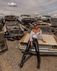 a woman sitting on the hood of a car in a junk yard full of old cars