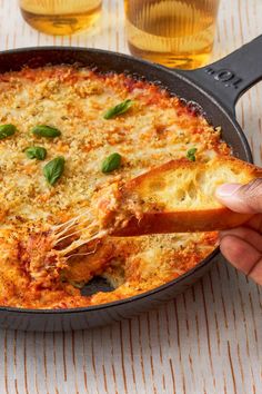 a hand holding a piece of bread over a skillet filled with lasagna