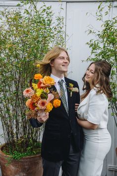 a man and woman standing next to each other in front of some plants holding flowers