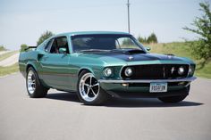 a green mustang sitting on the side of a road next to grass and trees in the background