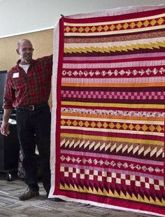 a man standing next to a red and yellow quilt in an office area with two people looking at it