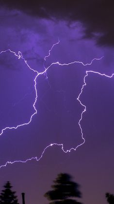 a purple sky with lightning striking through the clouds and some trees in front of it