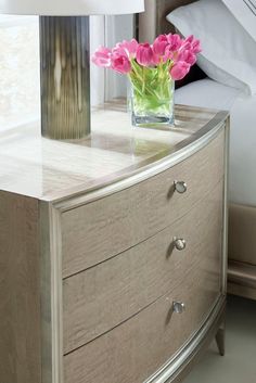 a vase with pink flowers sitting on top of a dresser next to a bed in a bedroom