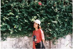 a woman with a hat is standing in front of a wall and some plants on the other side