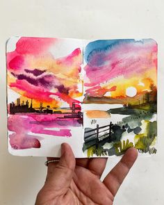 someone is holding up a watercolor book with an image of the sunset in it