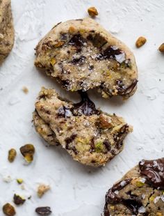 three cookies with chocolate and pistachio toppings on a white surface next to some pecans