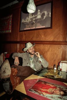 a man wearing a cowboy hat sitting at a table in front of a painting on the wall