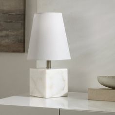 a white lamp sitting on top of a table next to a bowl and painting in the background