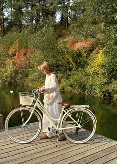 a woman standing next to a white bike on a wooden pier near water and trees