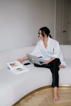 a woman sitting on top of a white couch holding a laptop next to an open book