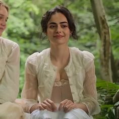 two women sitting next to each other in front of trees and bushes, one wearing a white dress