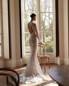 a woman standing in front of a window wearing a long sleeved wedding dress with an open back