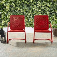 two red chairs sitting next to each other in front of a green wall with bushes