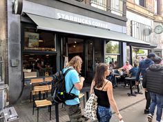 people walking down the sidewalk in front of a starbucks coffee shop with many tables and chairs