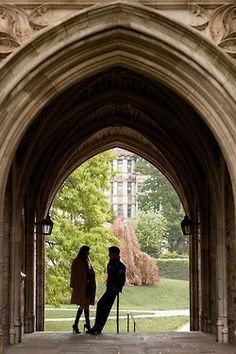 two people are standing under an archway in the middle of a campus with trees and buildings behind them